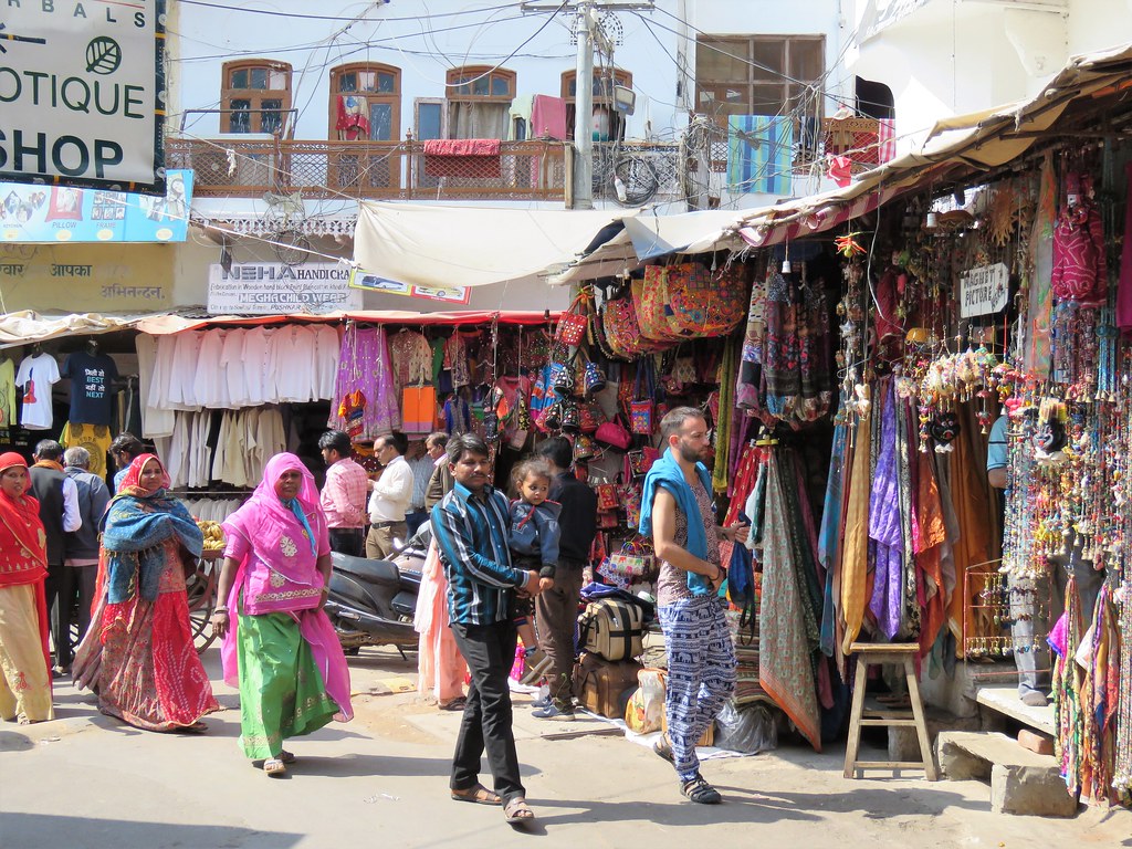 Women in bright Rajasthani saris walking through Pushkar bazaar, lined with textile and handicraft stalls
